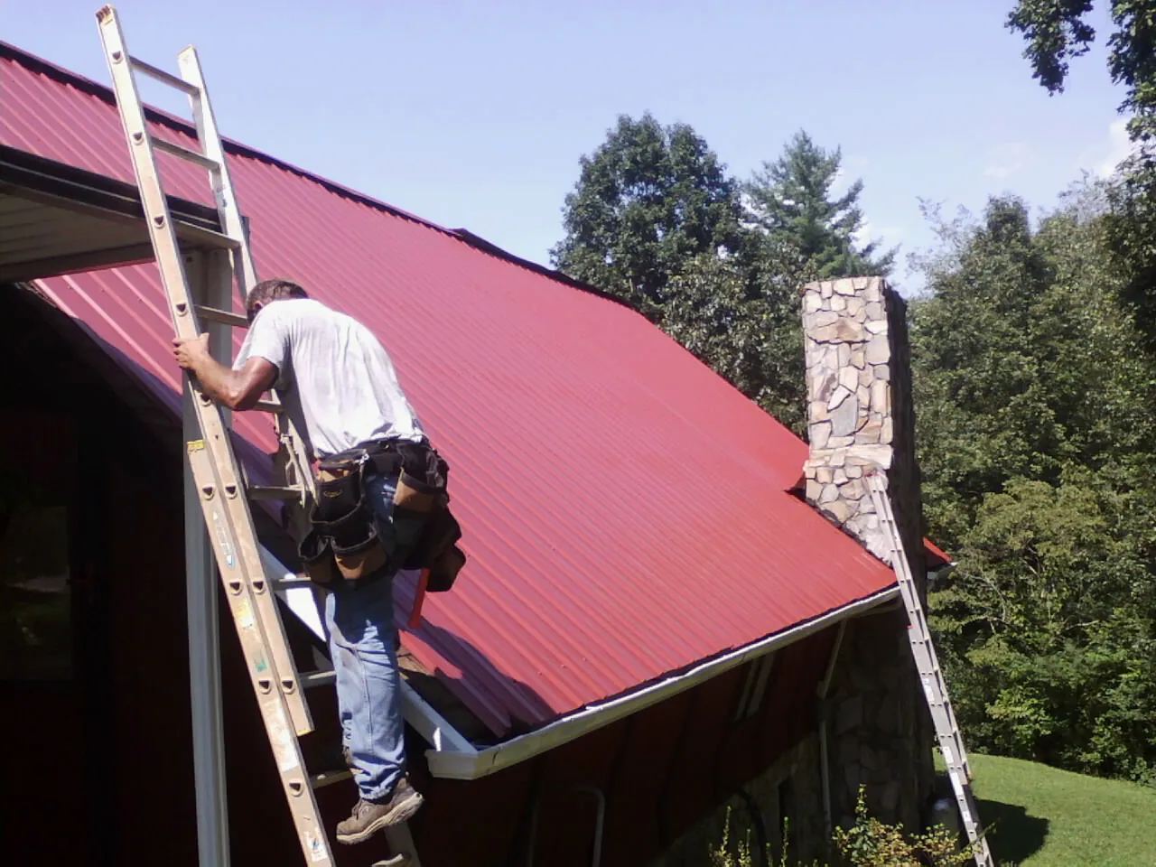 Roofer on a ladder beside a red metal roof and stone chimney — Soffit & Fascia Repair by Roofing Brothers in Hazleton